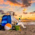 A man with an astronaut costume stands on the beach, wearing a backpack and holding a colorful beach ball, enjoying a sunny day by the ocean.