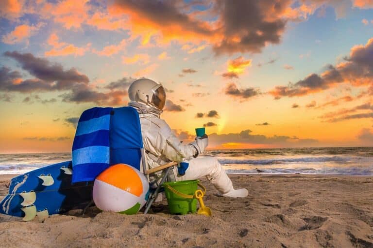 A man with an astronaut costume stands on the beach, wearing a backpack and holding a colorful beach ball, enjoying a sunny day by the ocean.